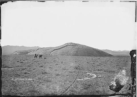 treeless plain with a low granite hill and two people riding on horseback in the distance