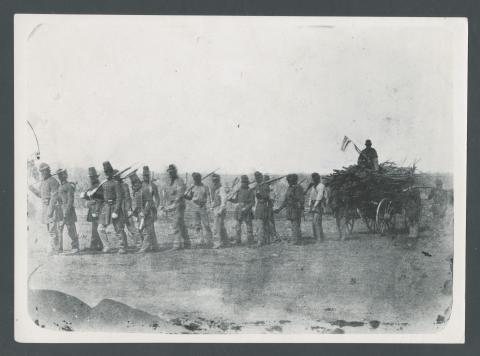 A wagon loaded with wood follows a line of uniformed soldiers carrying rifles in theri shoulder