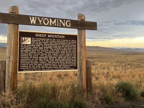 roadside sign hung from a wood crossbar that says, "Wyoming;" text on sign is titled "Sheep Mountain" and describes the geology of the areay