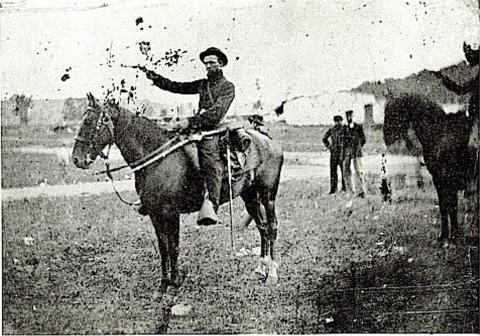 A uniformed man on horseback points his right arm forward as one other person on horseback and two men standing beyond him look on